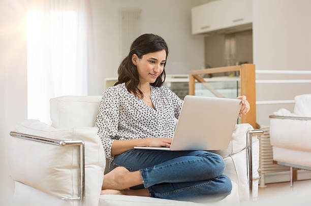 A woman sitting cross-legged in a white chair at home using a laptop.
