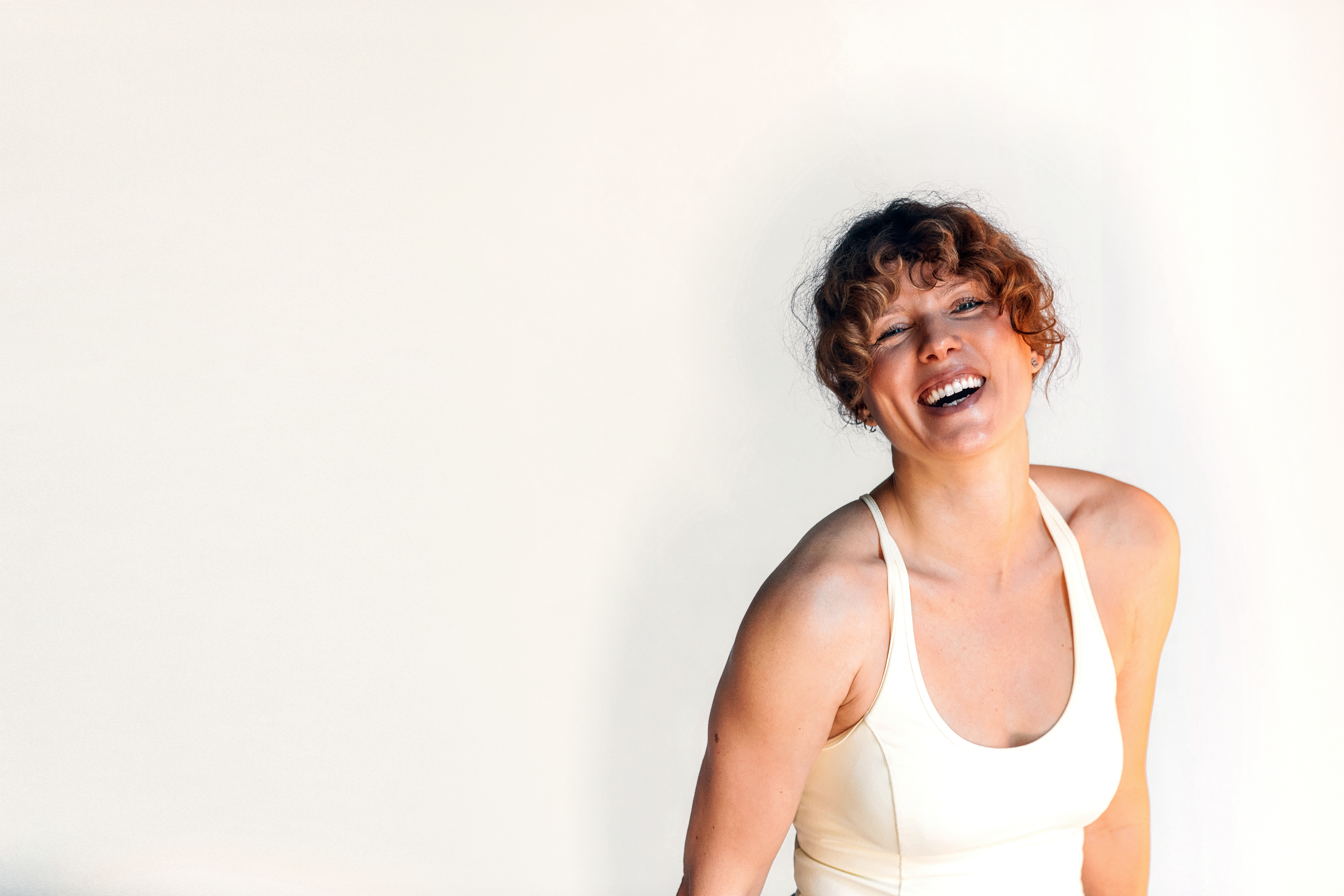 A smiling curly-haired woman in light activewear against a warm, bright background.